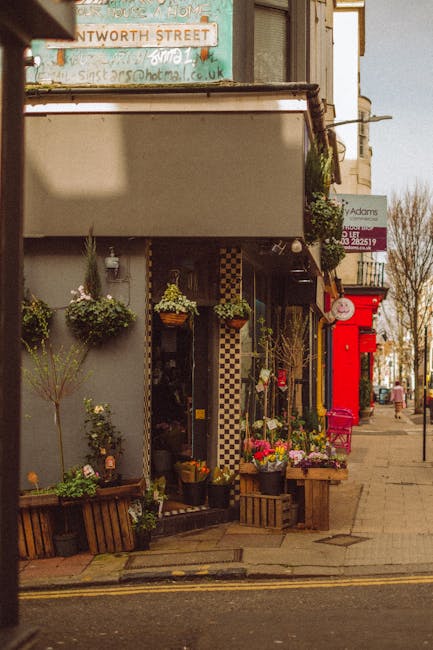 A storefront of a flower shop on a busy street, featuring an outdoor display of vibrant floral arrangements and potted plants. The arrangement includes mixed bouquets of fresh flowers such as roses, t