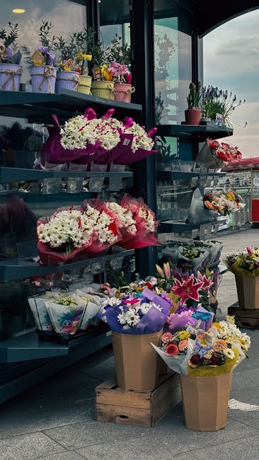 A display of vibrant floral arrangements and bouquets on dark gray shelves outside a flower shop, featuring a variety of fresh flowers including white chrysanthemums, pink lilies, and purple irises wr