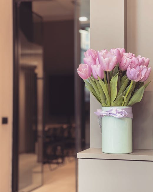 A bouquet of pink tulips with lush green leaves is arranged in a light green ceramic vase, which is decorated with a delicate white ribbon tied into a bow. The flowers are fresh, with gently curved pe