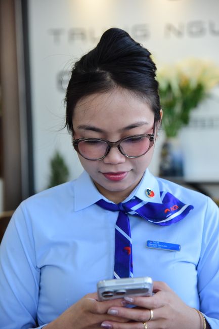 A close-up of a woman in a light blue uniform with a blue necktie, wearing glasses, holding a smartphone. Behind her, there is a bouquet of white roses with green leaves arranged in a vase, placed on