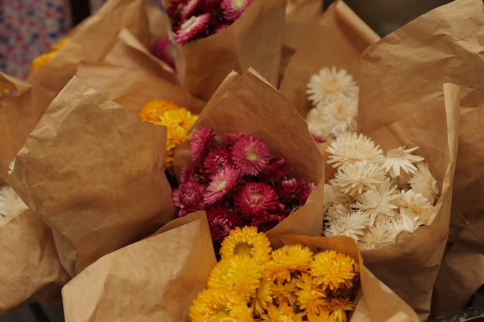 A selection of fresh flower bouquets wrapped in brown kraft paper, featuring vibrant pink chrysanthemums, bright yellow daisies, and soft white daisies. The flowers are arranged in small, individual b