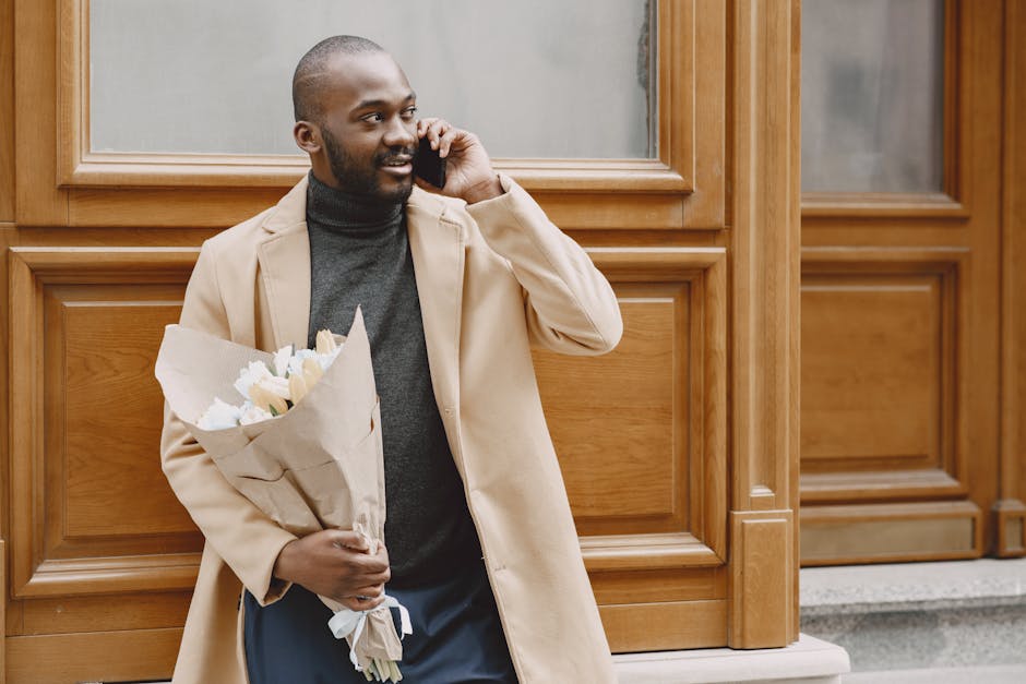 A man with dark skin, a beard, and a bald head is standing outdoors in front of wooden paneling, holding a bouquet of fresh white roses wrapped in brown paper. He is wearing a beige coat over a dark g