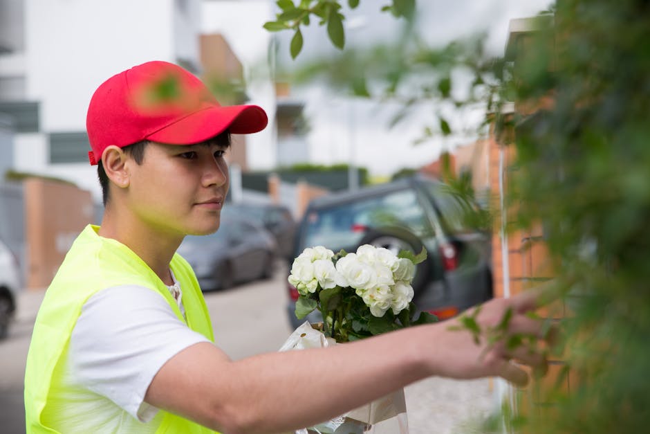 A young male florist delivering a floral gift in an outdoor residential area. He is wearing a red cap, a white t-shirt, and a yellow high-visibility vest, and is holding a bouquet of white hydrangeas